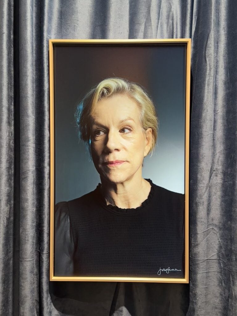 a middle aged white woman's face is framed by a gold frame and gray curtains