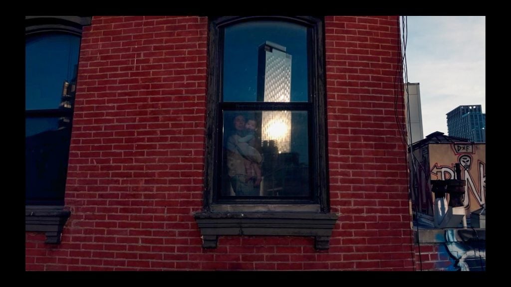 Carissa Rodriguez, Mother and Baby. A film still of a woman and her child seen through the window of a brick apartment building.