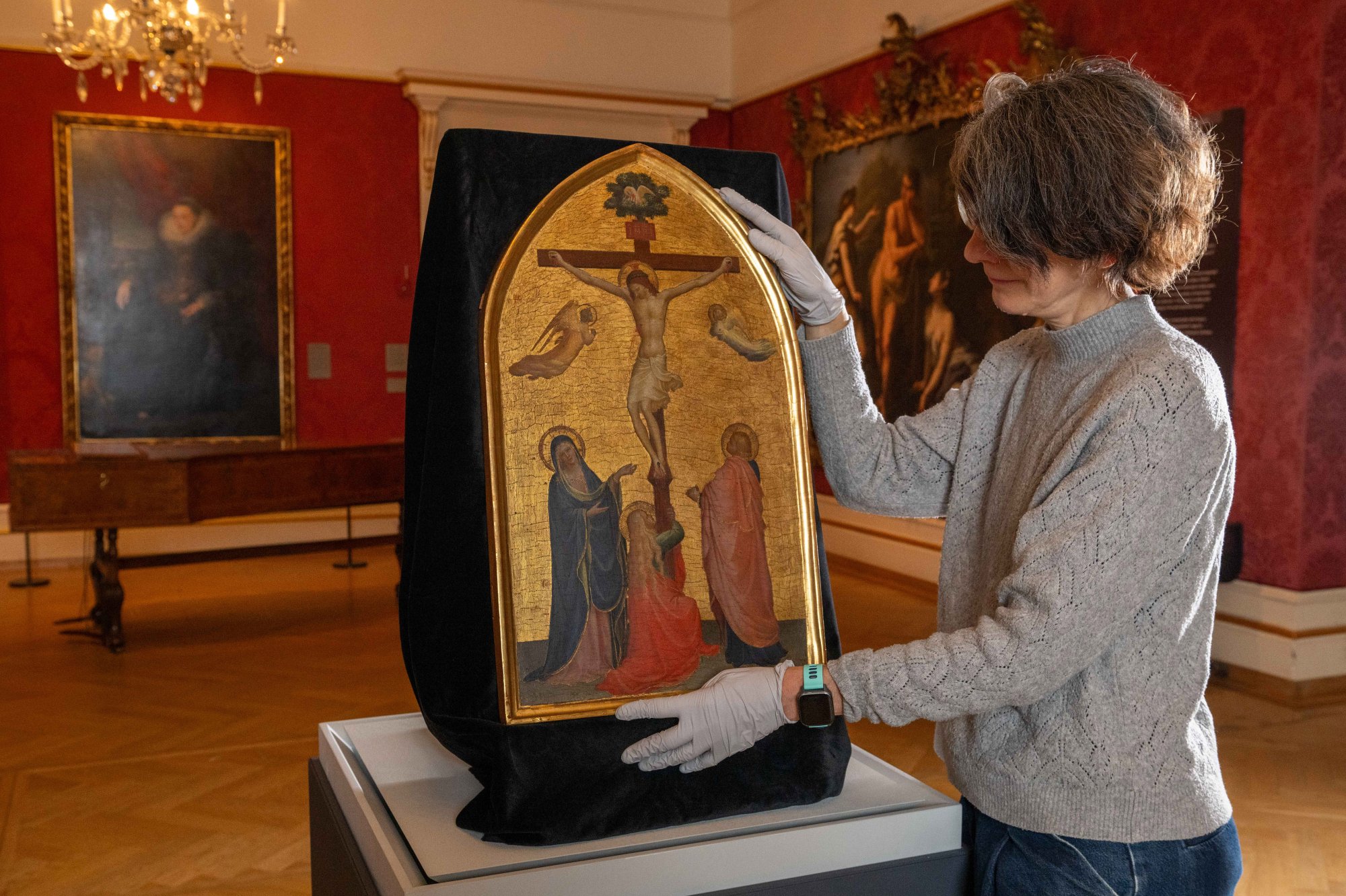 a woman in a red walled museum interior covered in old paintings can be seen in a grey cardigan and in profile, she uses gloves to hold the edges of a medieval gold painting with a cruxifion scene on a gold ground and devoted figures standing around beneath Jesus on the Cross