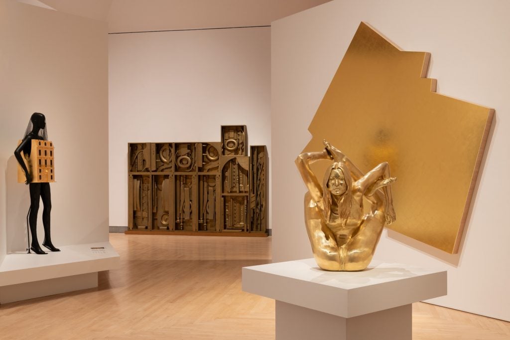 a museum installation depicts a contorted gold-plated statue and other golden works