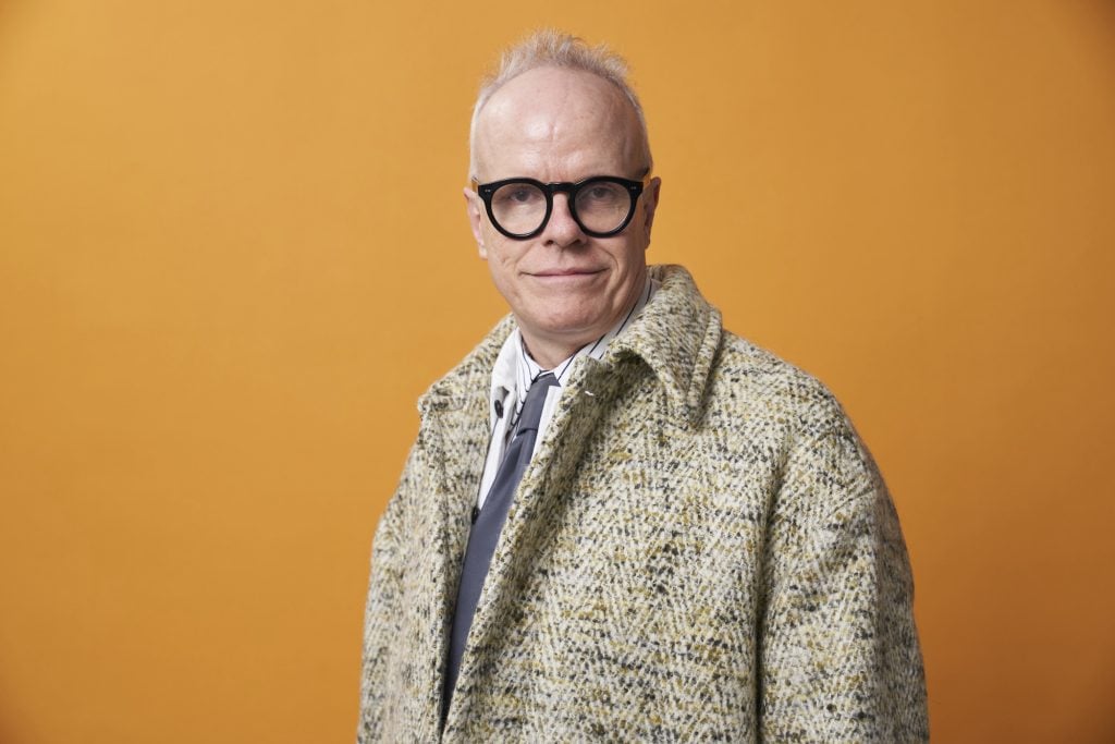 Hans Ulrch Obrist wearing glasses and a tan coat stands in front of an orange background