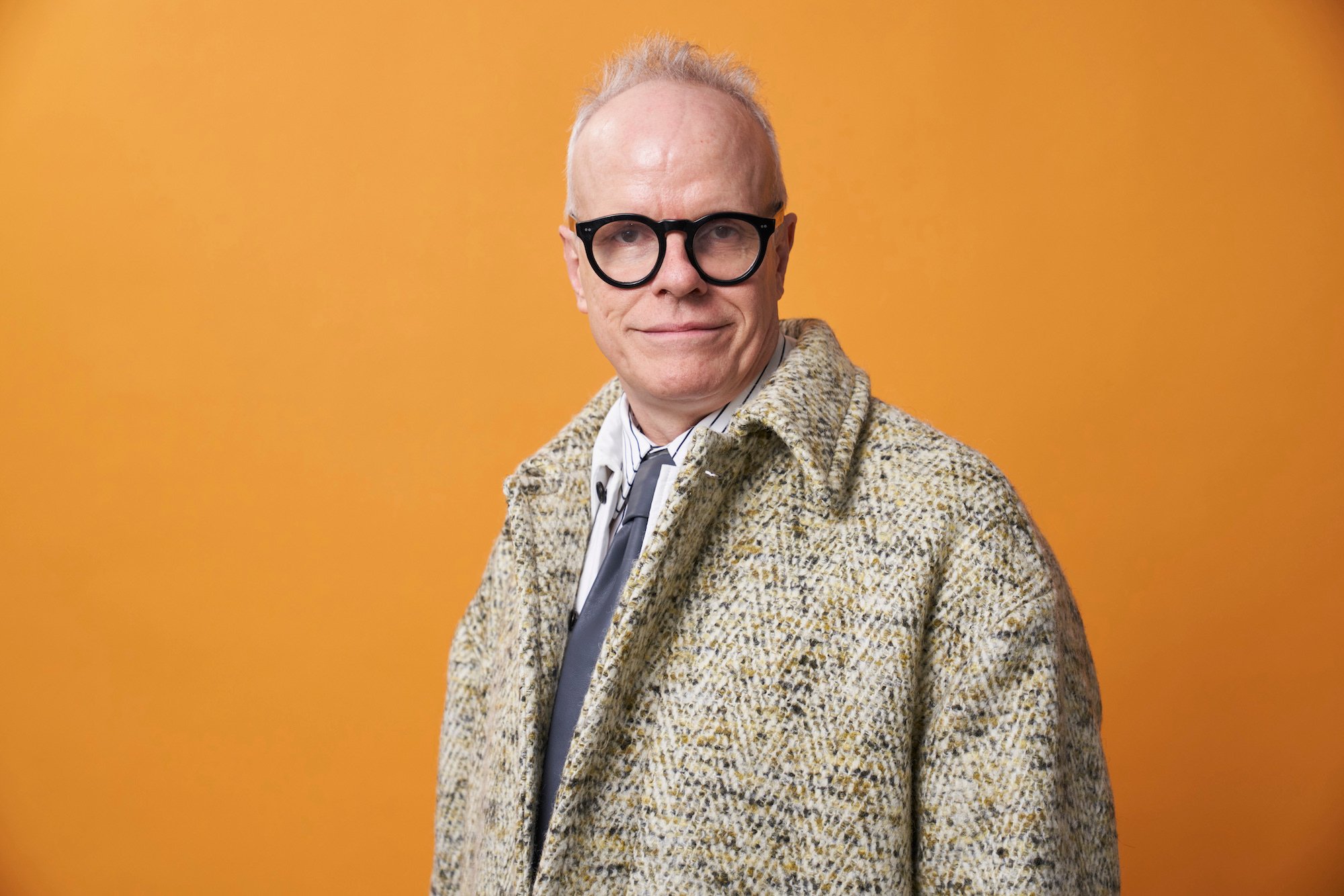 Hans Ulrch Obrist wearing glasses and a tan coat stands in front of an orange background