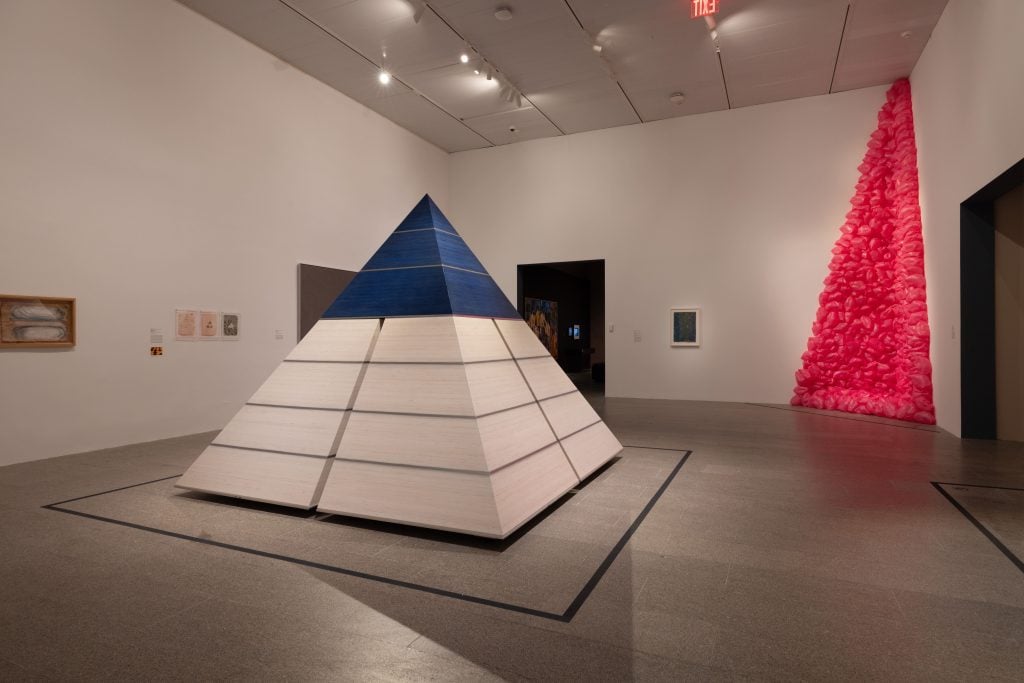 a museum gallery has a white wooden pyramid and an abstract pink plastic one 