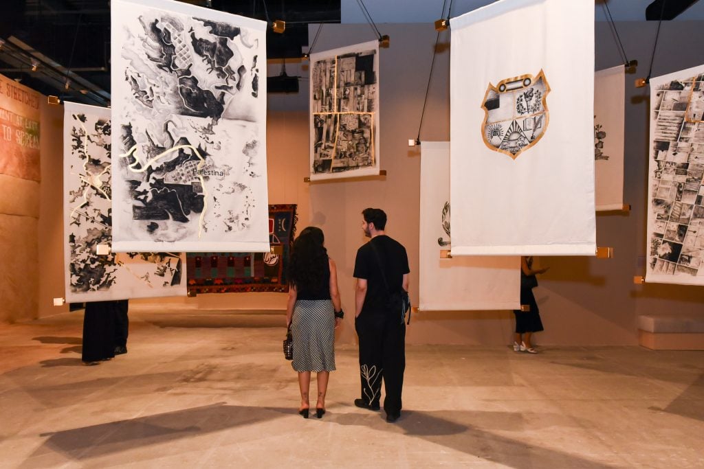 Two visitors observe an art installation featuring large, suspended fabric panels with intricate black-and-white designs, maps, and crests in a gallery setting.