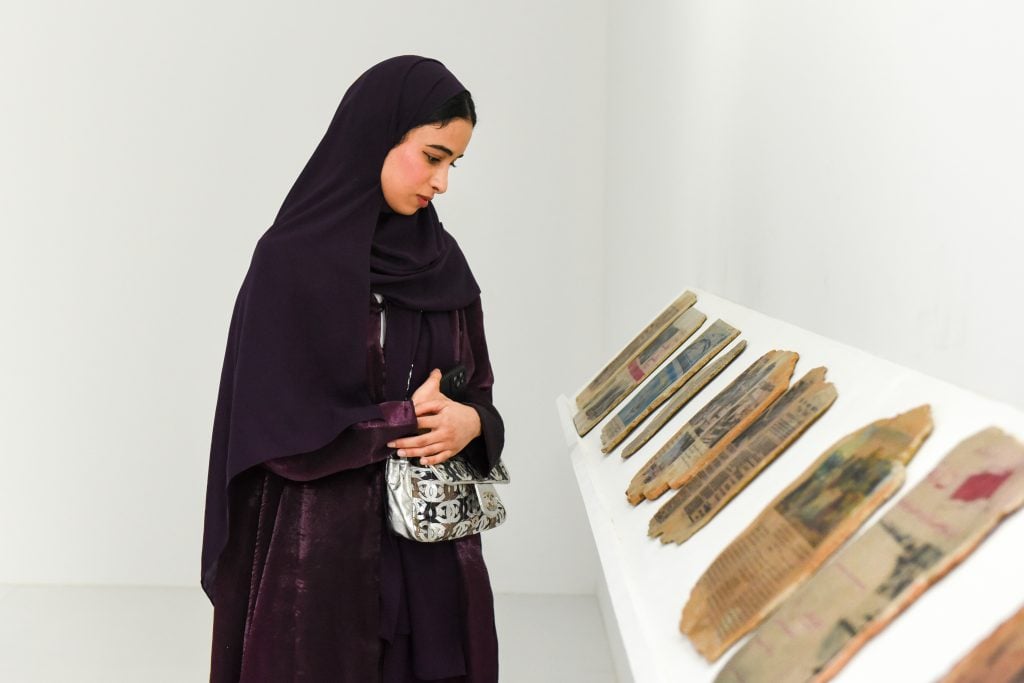 A woman wearing a deep purple abaya observes a display of aged, fragmented newspaper pieces on a white slanted platform in an exhibition.