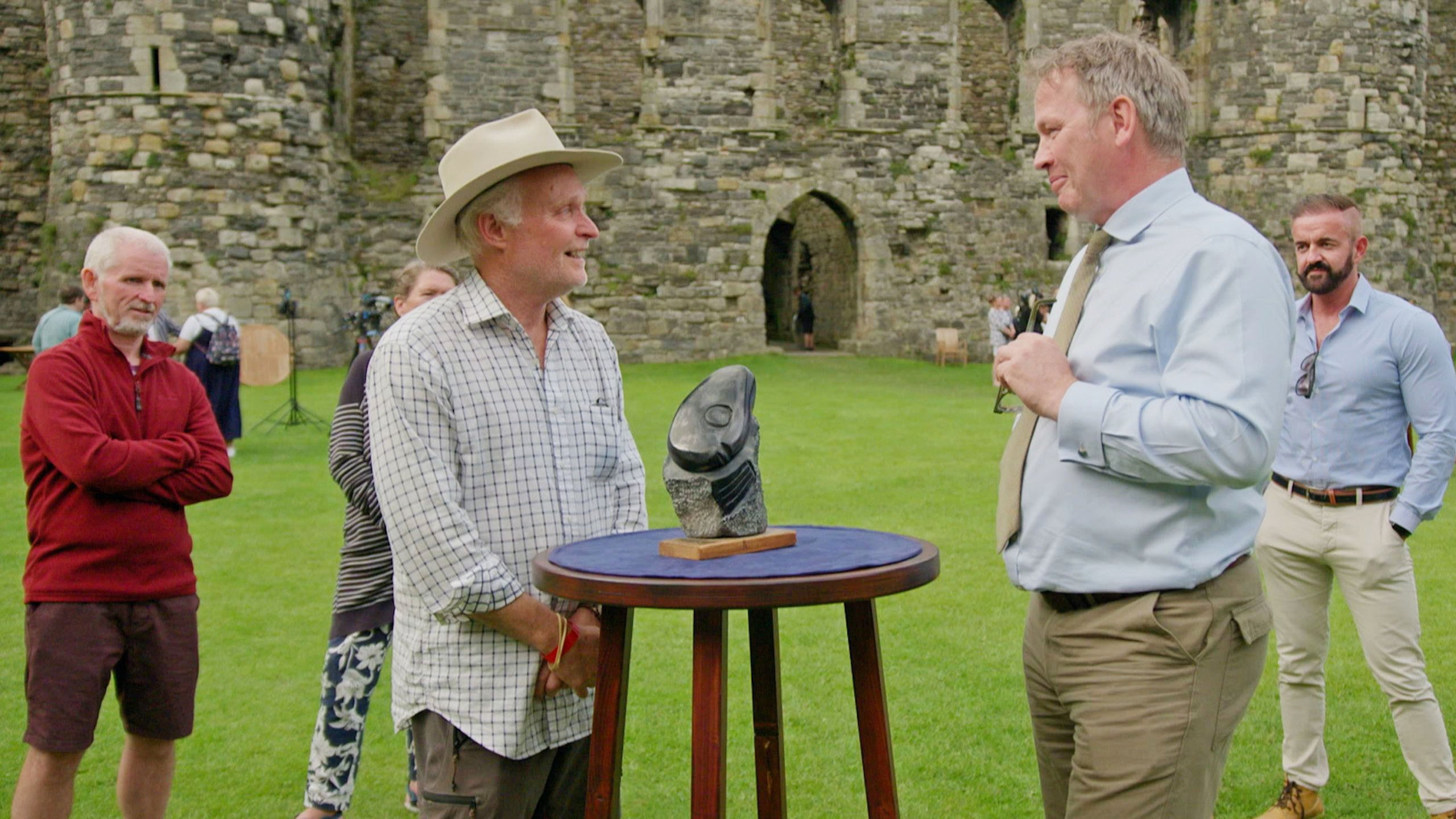 Two men stood on grass in front of an ancient castle door looking at a stone sculpture on a raised wooden table