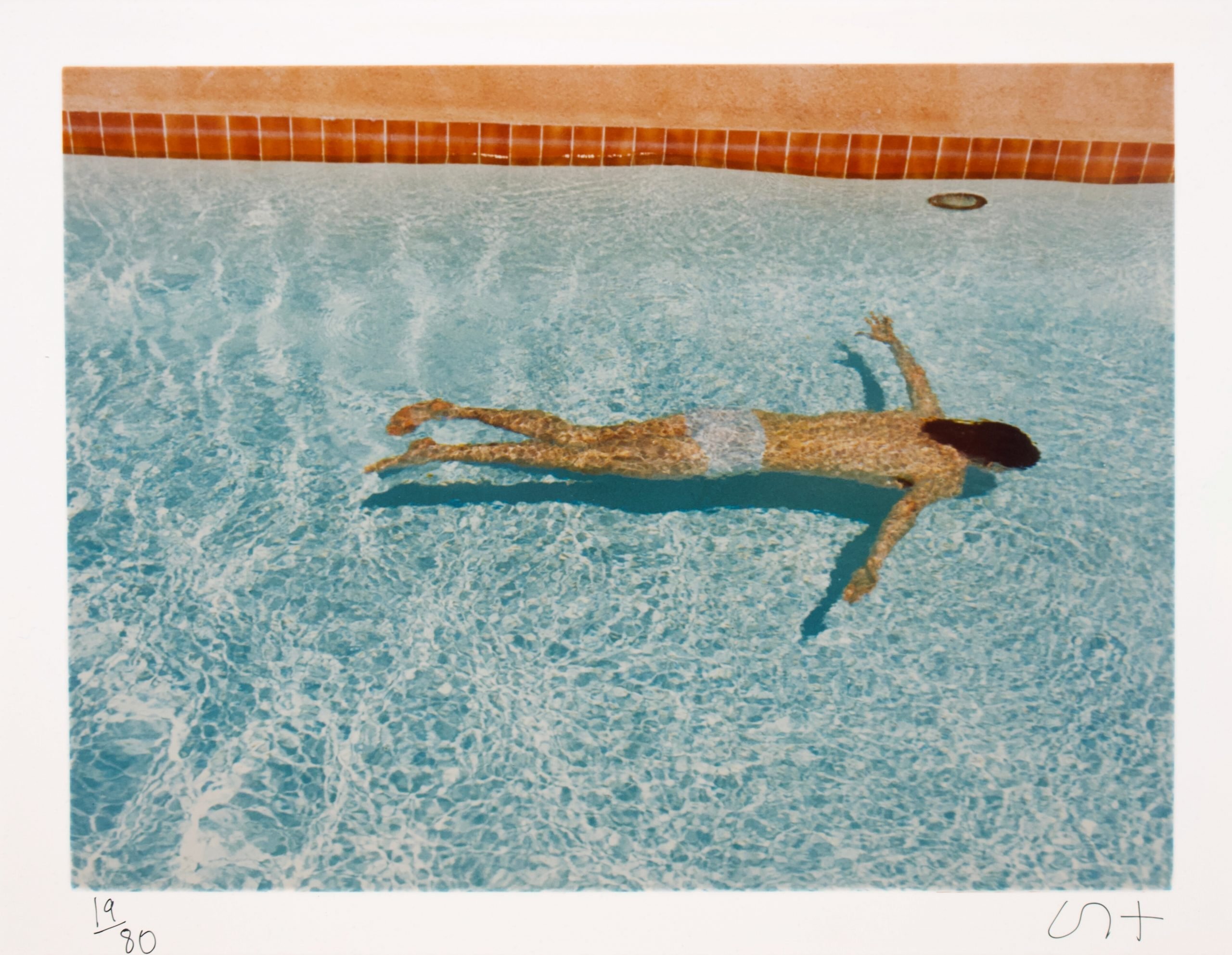 A person floats face-down in a sunlit swimming pool, surrounded by clear blue water, casting a shadow on the pool floor.