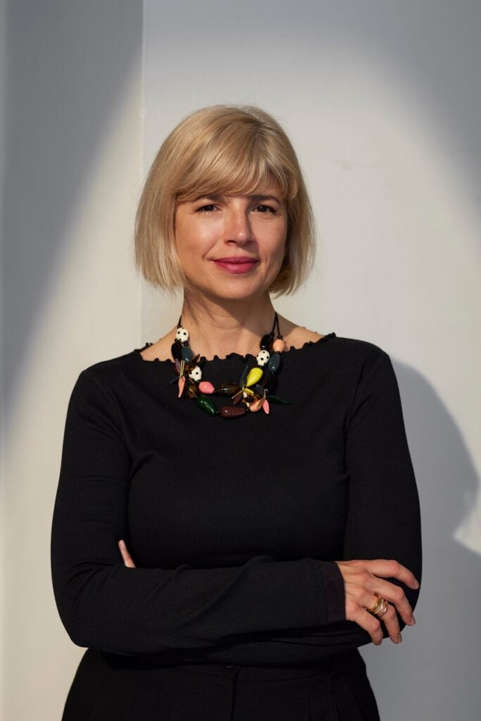 a woman with blonde hair and a black dress stands against a white wall with shadows