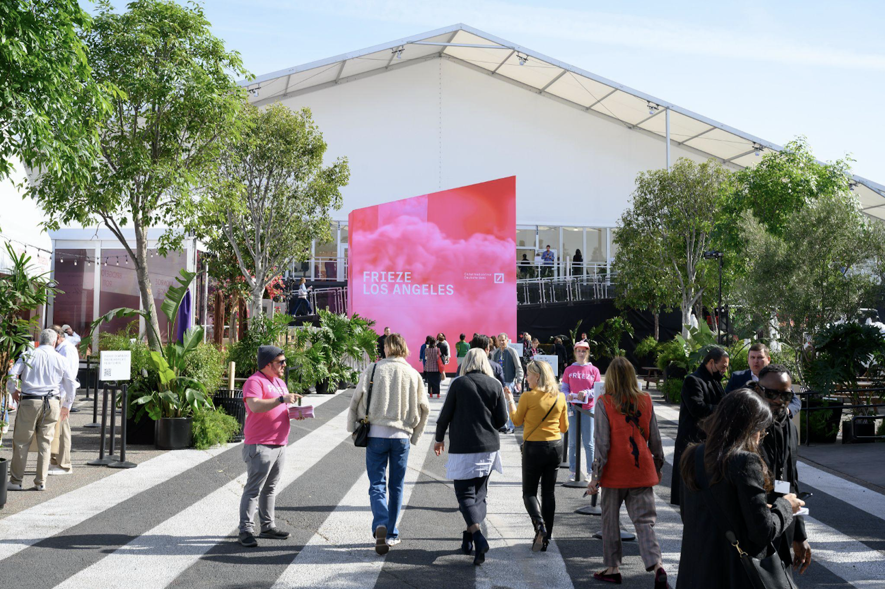 a crowd of people walks toward a large white outdoor tent. a pink sign reads frieze los angeles