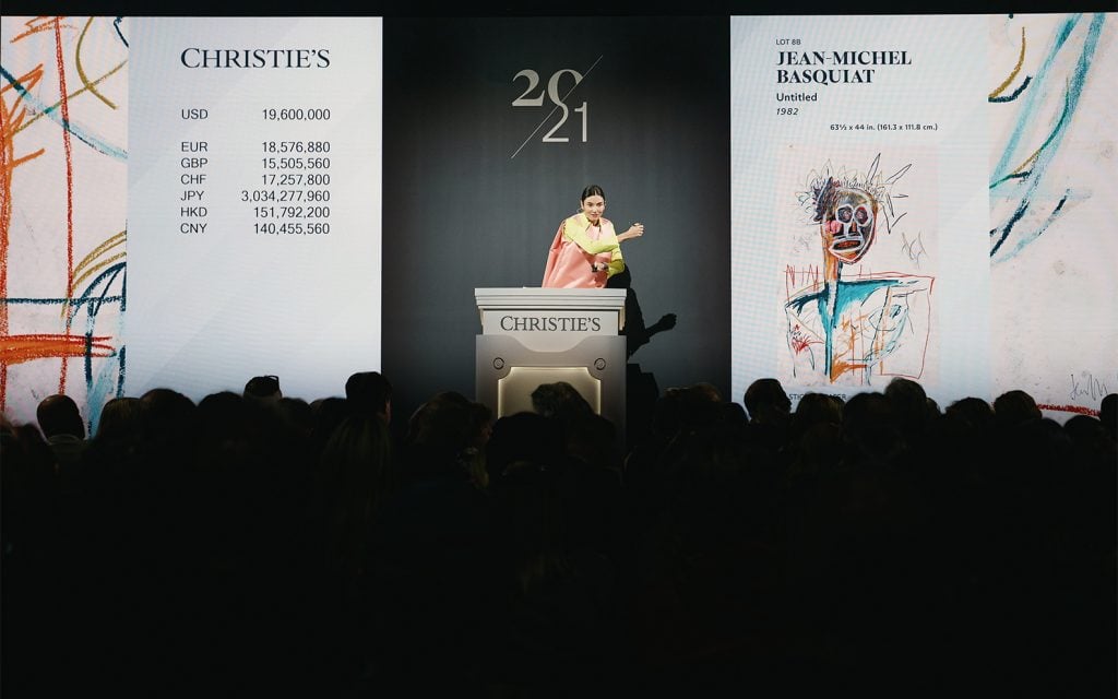 A Christie’s auctioneer stands at the podium, selling a Jean-Michel Basquiat artwork displayed on a screen, with prices listed in multiple currencies.