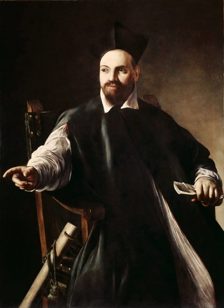 a man in a black throw sits in a chair with a finger outstretched