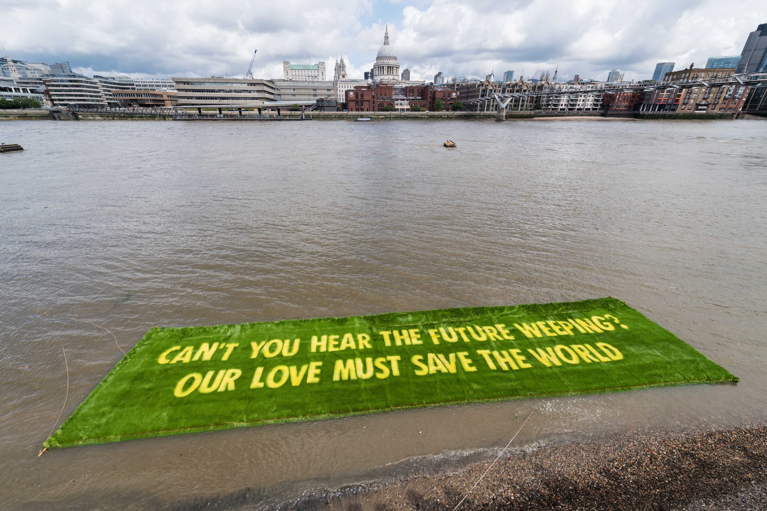 A large green floating installation on the Thames with the words, "CAN'T YOU HEAR THE FUTURE WEEPING? OUR LOVE MUST SAVE THE WORLD," against London's skyline.