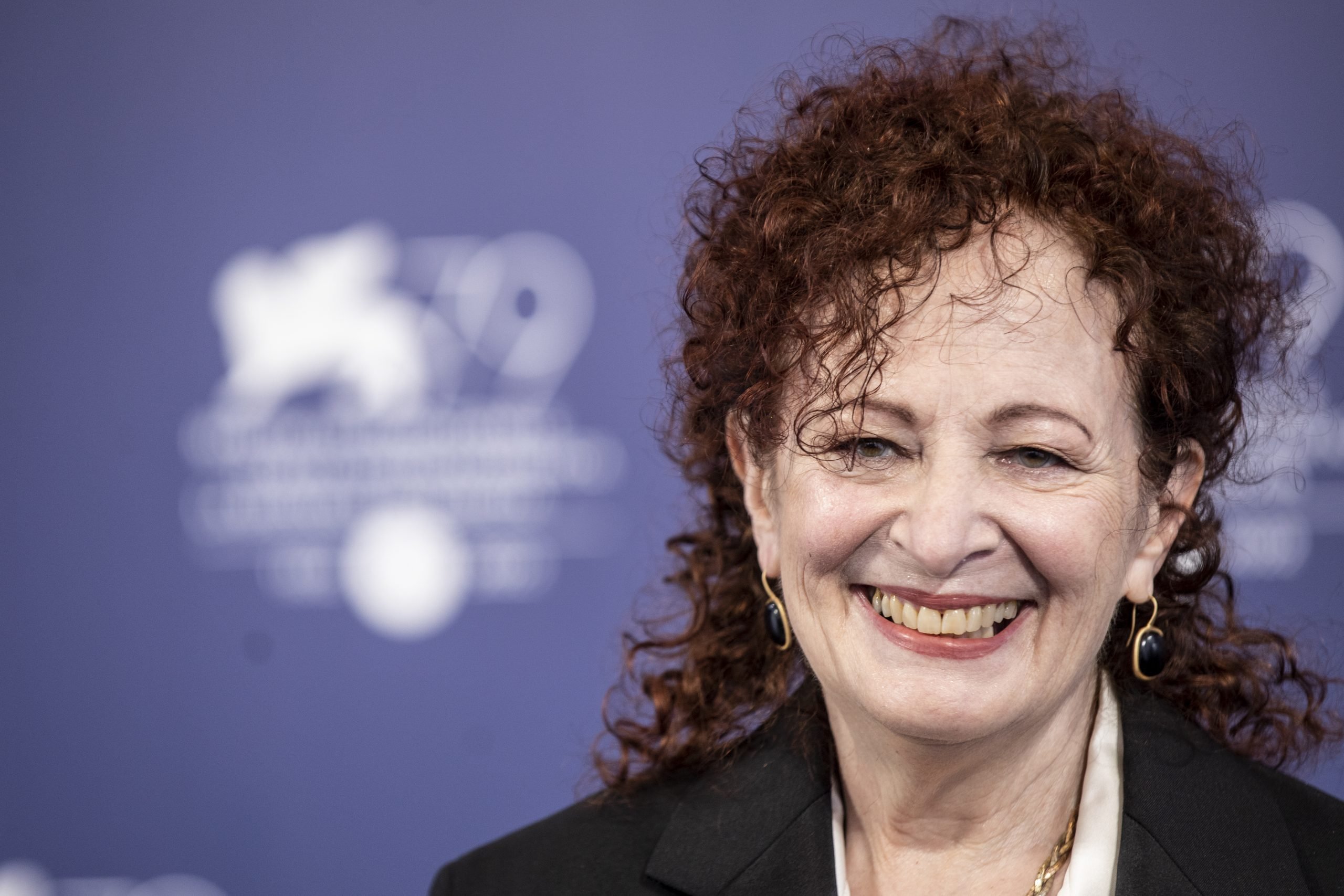 a woman with dark red curly hair smiles at the camera in front of a purple backdrop