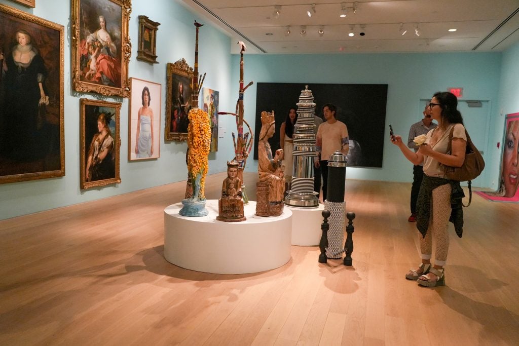 The Bass Museum of Art in Miami Beach in 2022. The gallery is hung salon style, with a circular platform in the center of the room featuring several totemic sculptures. One of the museum goers is taking a photograph with her phone. 