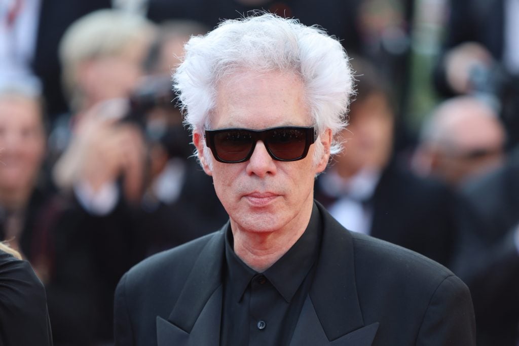 a man with white hair and black sunglasses in a black suit is seen in front of a crowd