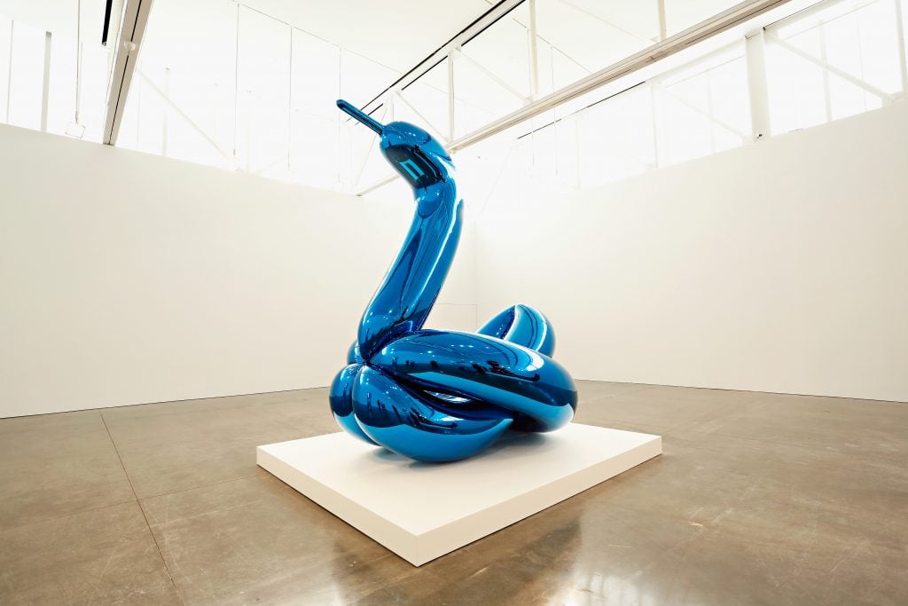 a large blue sculpture in the shape of a balloon animal swan stands alone in a large white gallery