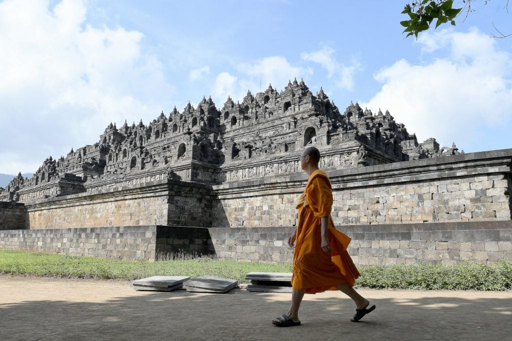 A Buddhist monk in orange robes walks by the ancient Borobudur Temple in Indonesia, set against a bright, partly cloudy sky.