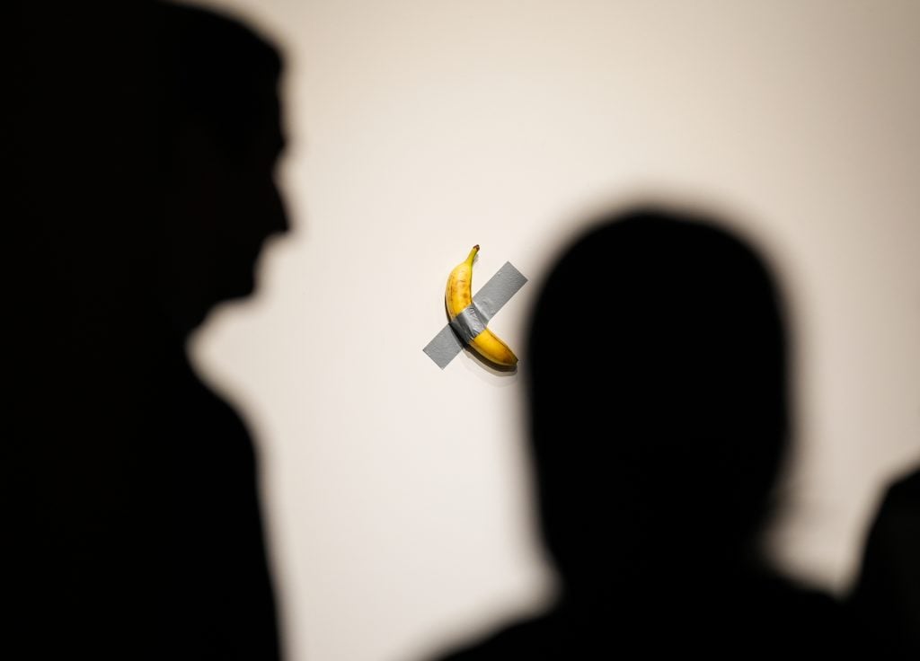 the shadowed backs of two people look at a banana taped to the wall