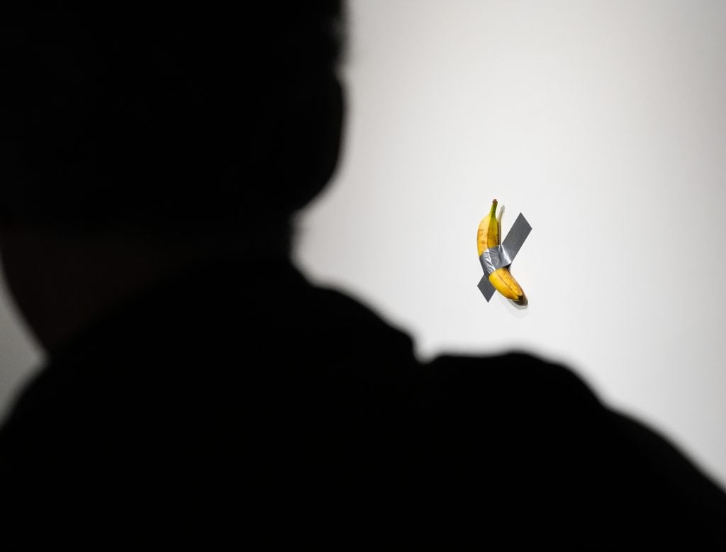 A silhouetted person observes a banana duct-taped to a white wall