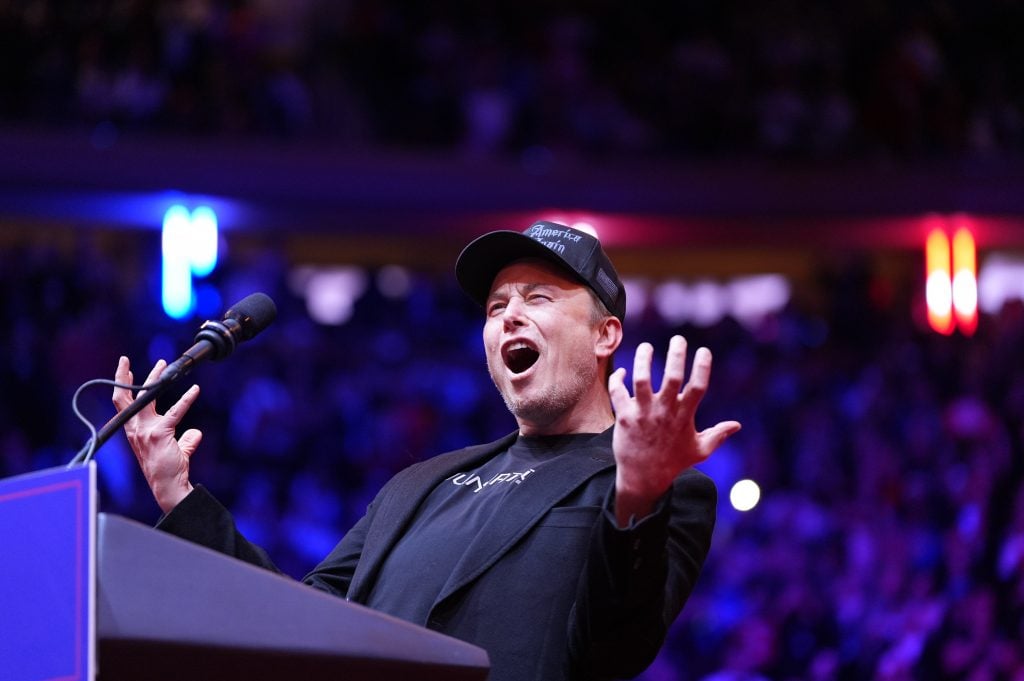  A man in a black cap and blazer passionately gestures while speaking at a podium, set against a colorful, blurred background of an audience