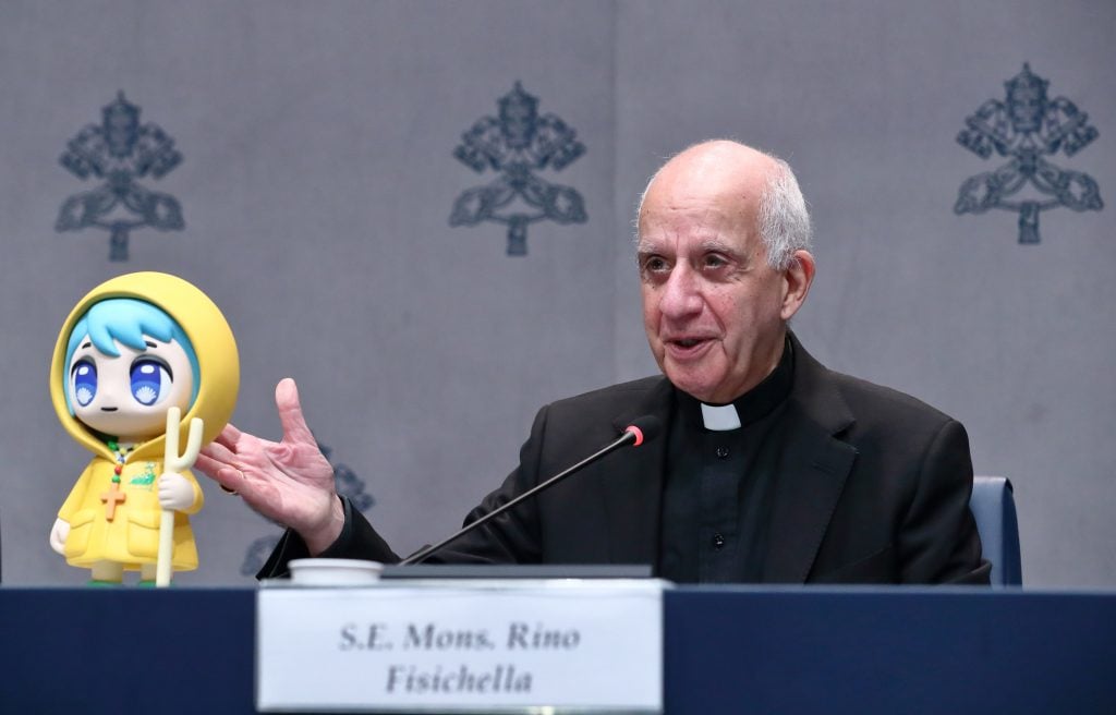 a older man in a black religious attire sits behind a guest with a microphone pointed at him, he is gesturing to a little cartoon figurine of a girl with blue hair in a yellow raincoat