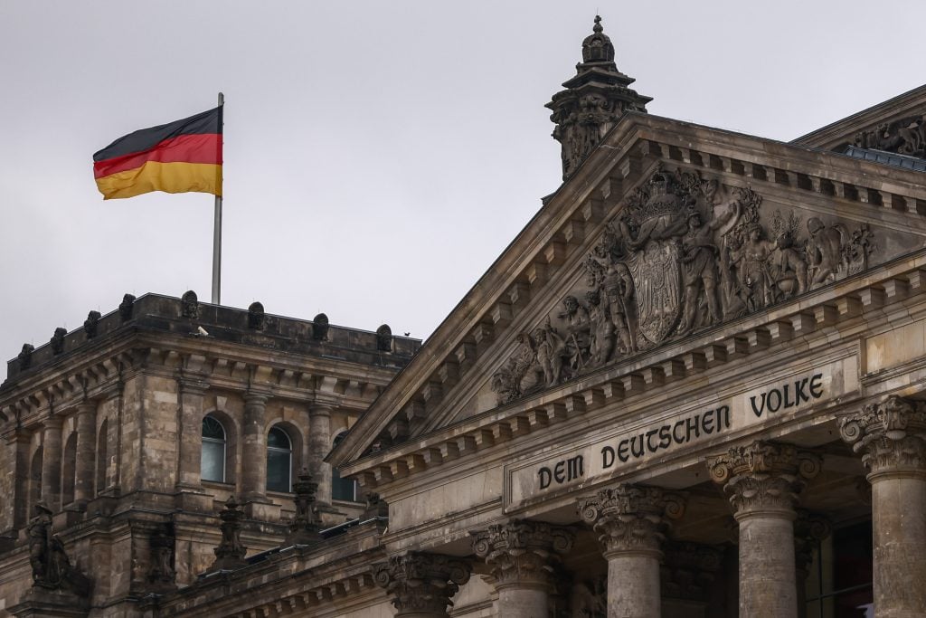 The German flag waves atop the historic Reichstag building in Berlin. The neoclassical facade features ornate sculptures and the inscription 