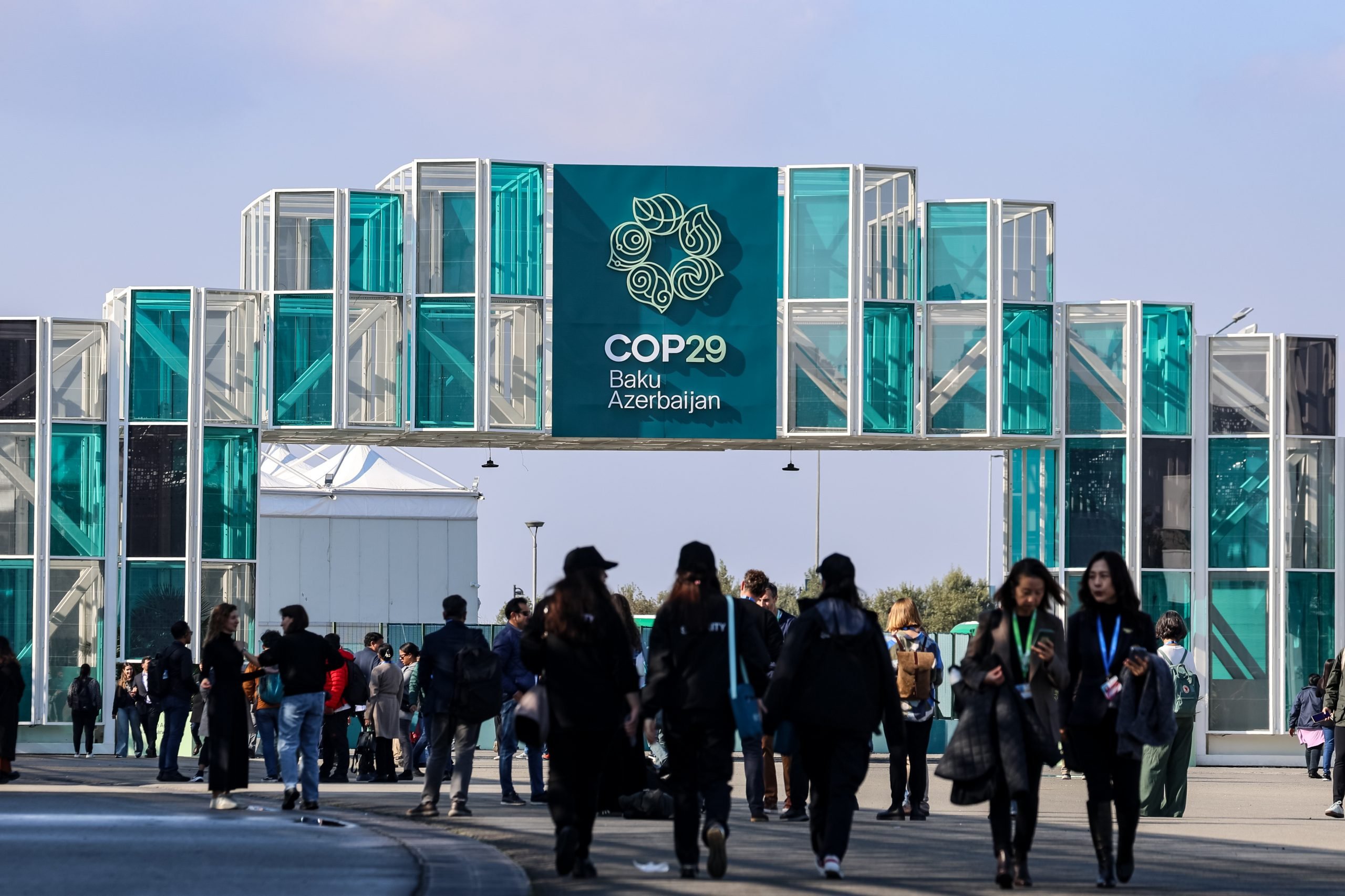 Crowd walking towards the entrance of COP29 in Baku, Azerbaijan. The modern structure displays a large event banner in teal.