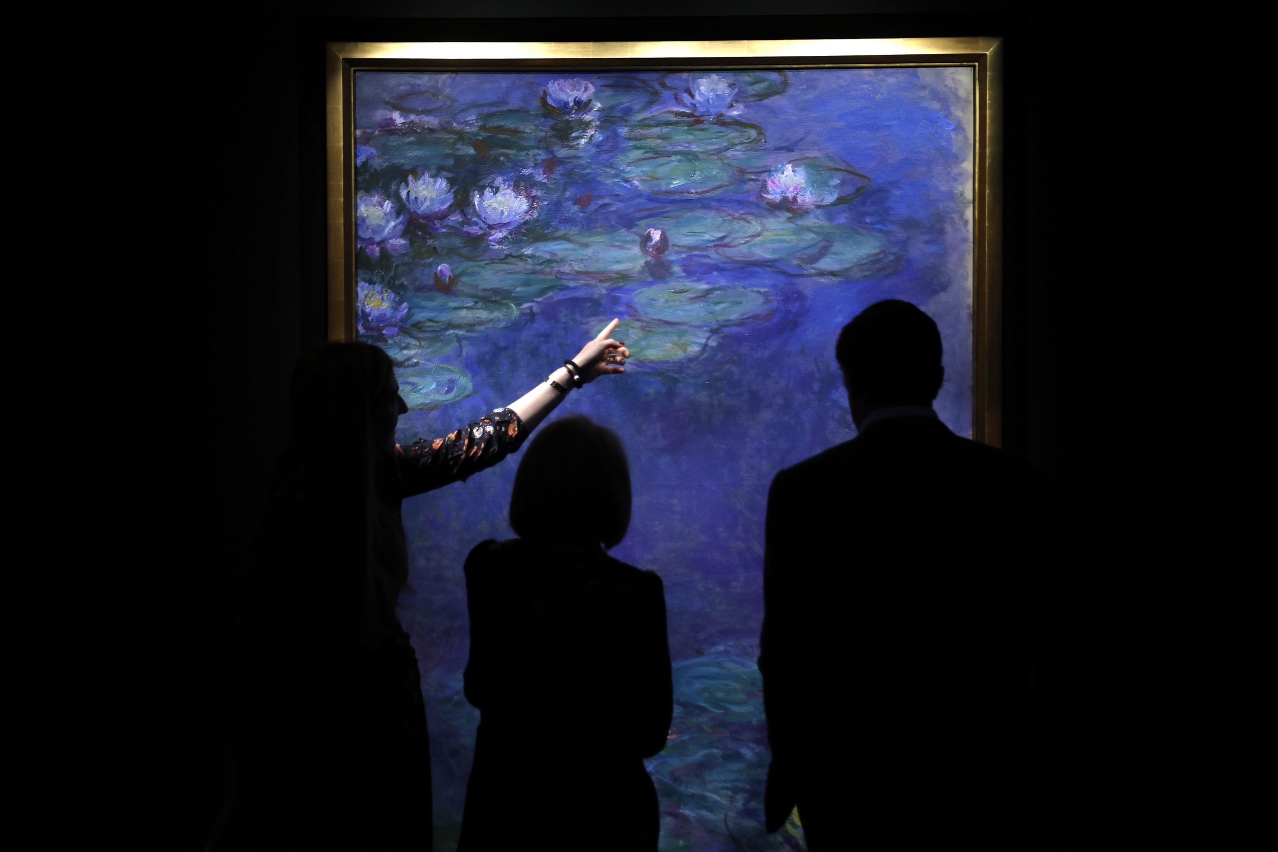 Silhouettes of three people are seen in front of a large framed painting of Claude Monet's water lilies. One person gestures towards the vibrant blue and green artwork, highlighting its details in a dimly lit gallery space.