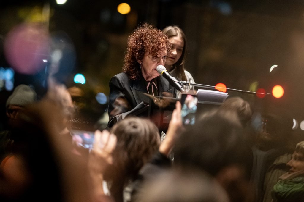 A woman with curly hair speaks into a microphone at an evening event, surrounded by an attentive crowd holding up phones to record.