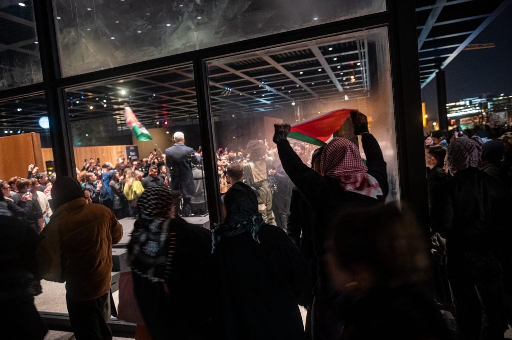 A crowd gathers outside a glass-walled venue at night. People inside hold flags and record the event, while others watch and wave flags outside.