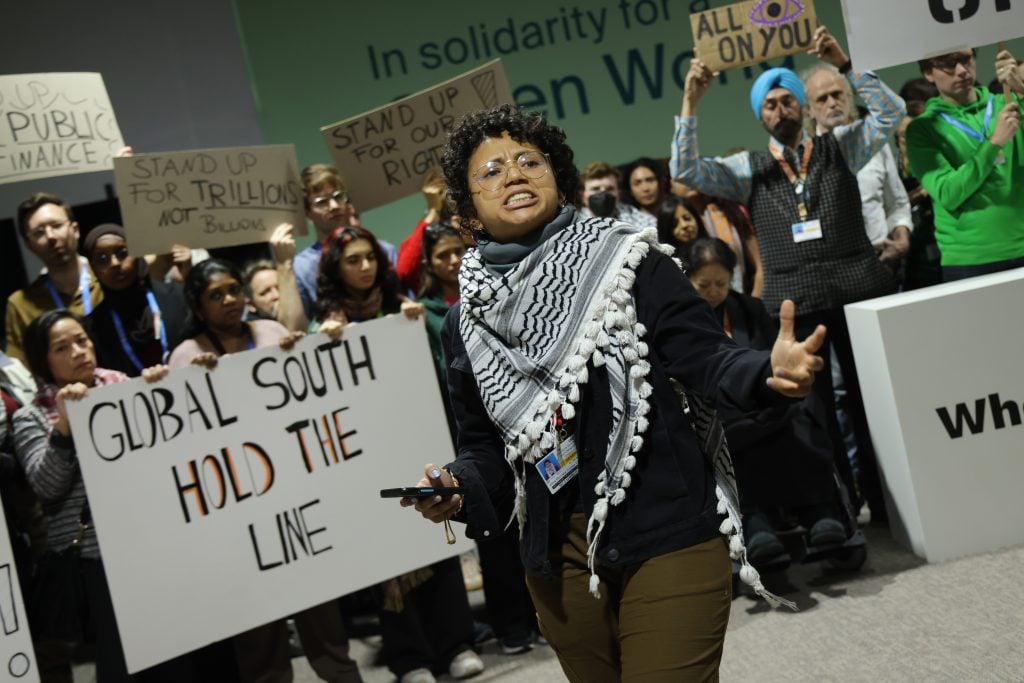 A passionate activist speaking at a climate protest, surrounded by diverse demonstrators holding signs, including "Global South Hold the Line" and "Stand Up for Our Rights."