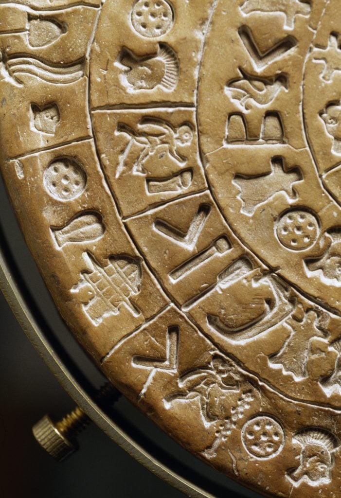 a close up of the Phaistos Disk, revealing the hieroglyphs which include a walking man, a dotted shield, a fish, and other decipherable figures.