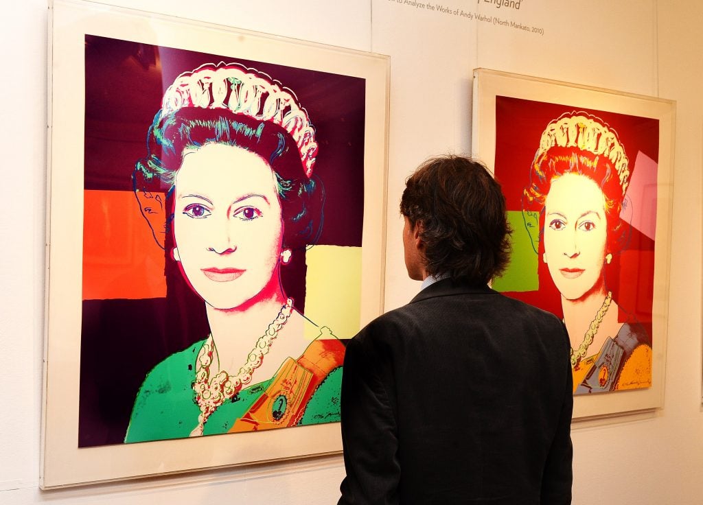 A man observes vibrant, pop art portraits of Queen Elizabeth II, rendered in bold colors and contrasting backgrounds, reminiscent of Andy Warhol’s style.