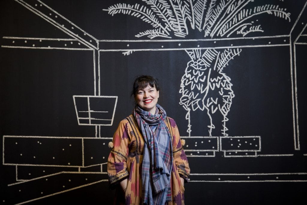 A smiling woman stands in front of a black wall with white chalk-style drawings of palm trees and geometric designs, wearing a colorful outfit.