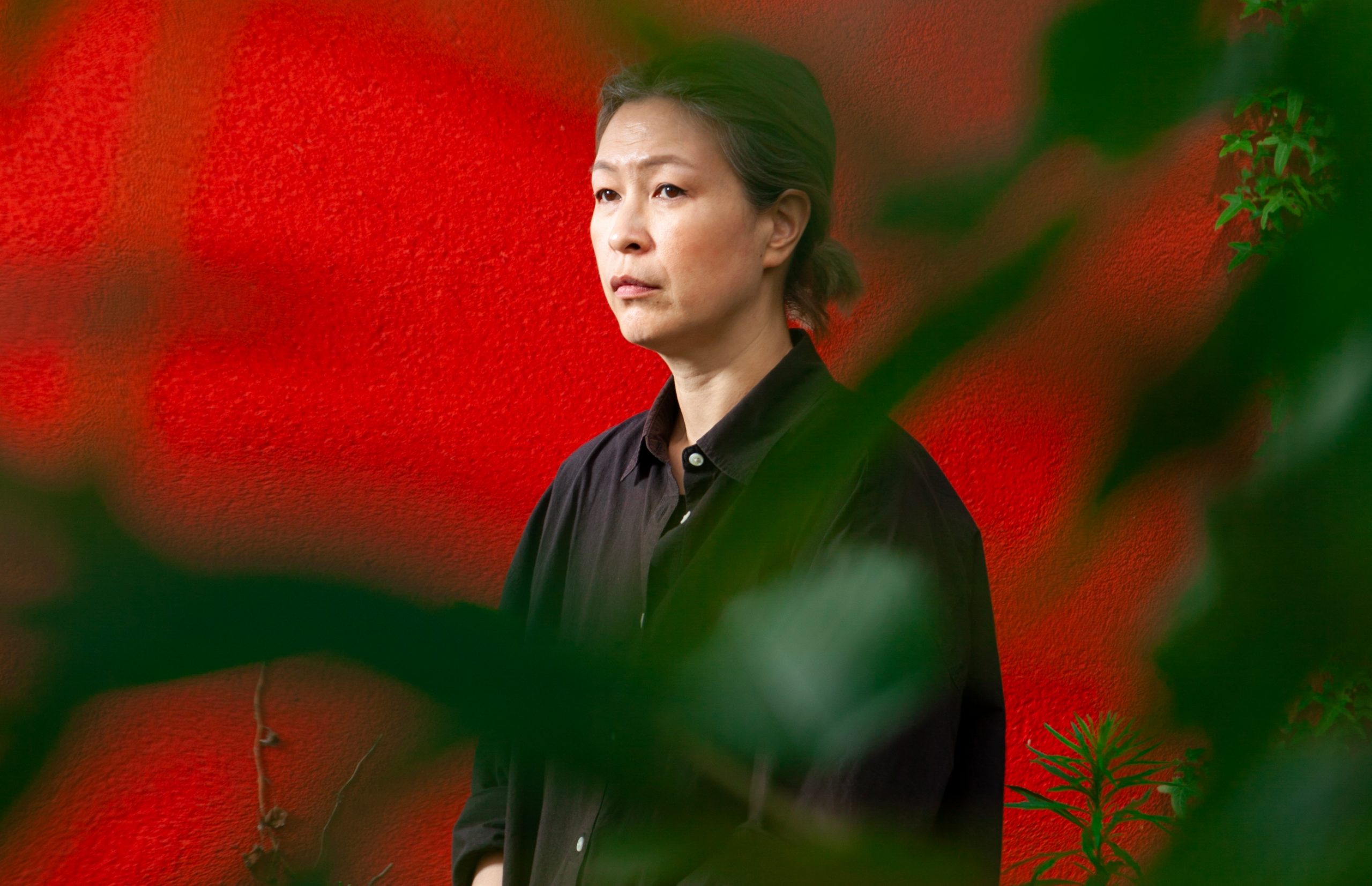 a woman in a black shirt stands against a bright red wall and is seen through tree leaves in the foreground