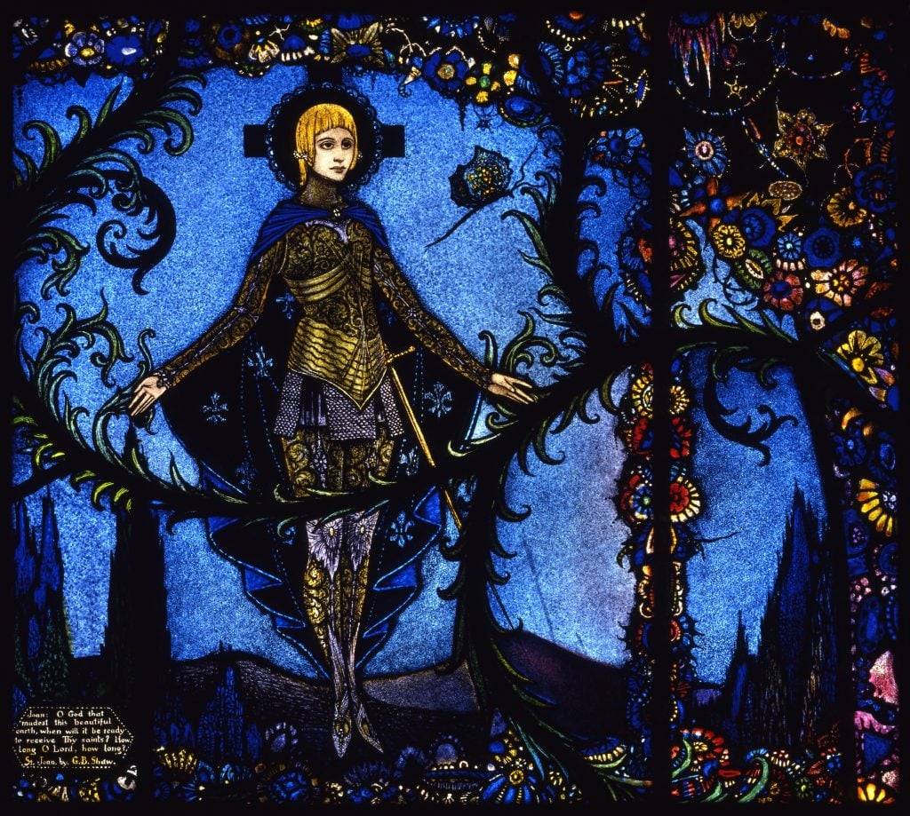 Harry Clarke, St. Joan by George Bernard Shaw from Harry Clarke Geneva Window (1926–30). A beautifully rendered stained glass window pane with St. Joan of Arc in her armor against a blue background with colorful flowers and vines. 