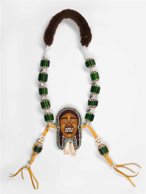 A handcrafted Native American-style necklace featuring green and white beads strung along with braided yellow leather cords. The centerpiece is an intricate beadwork medallion depicting a face with prominent white teeth, adorned with turquoise accents and hanging shell or bone ornaments. The necklace also includes fur padding at the top.