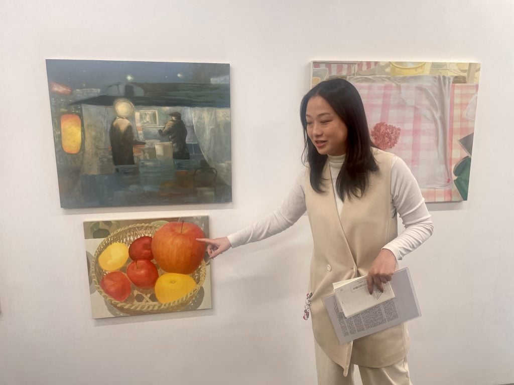 An East Asian woman in long hair, beige clothes, talks about the three paintings hung on the white wall behind her. She is pointing at a painting full of fruits. 