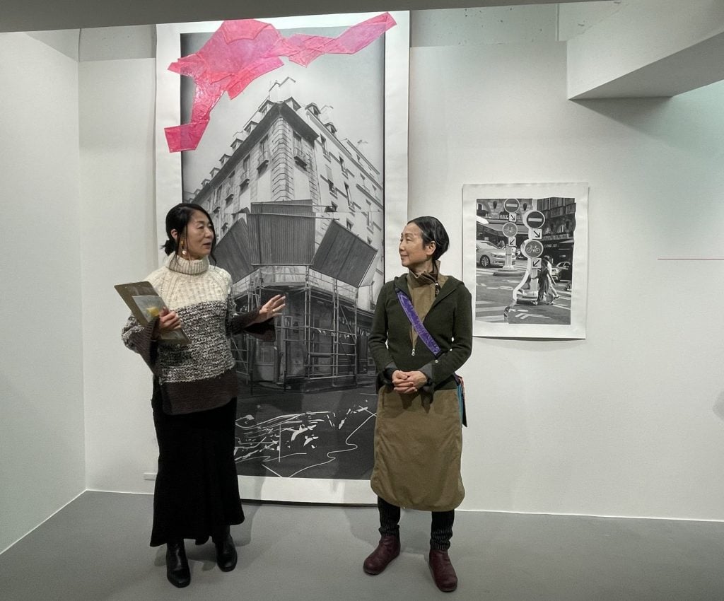 Two East Asian women standing in front of a large black and white photography work with a splash of pink paint.