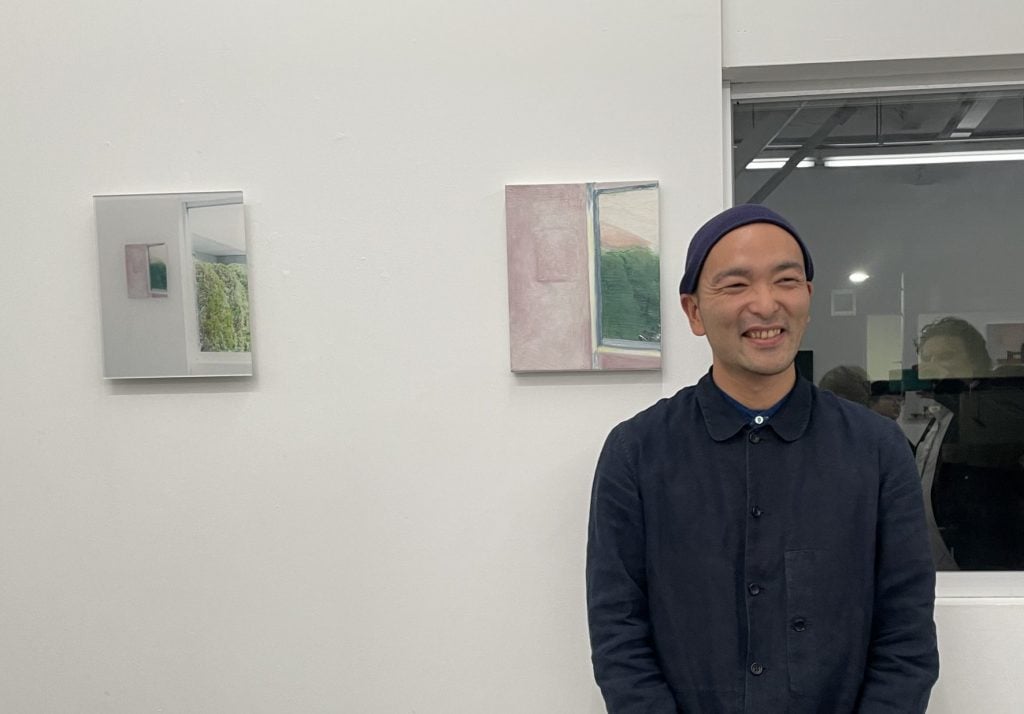 An East Asian man in a beanie dressed in blue smiling. He is standing next to one painting and a photo hung on a white wall. 