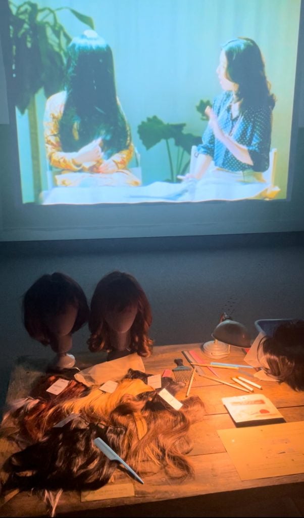 A large video screen on the wall showing a woman journalist in blue blouse interviewing another being whose face is covered by long hair. In front of the screen is a table that sits various types of wigs.