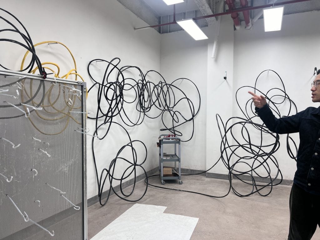 An artist in his studio with his work, which resembles looping, intertwined wires