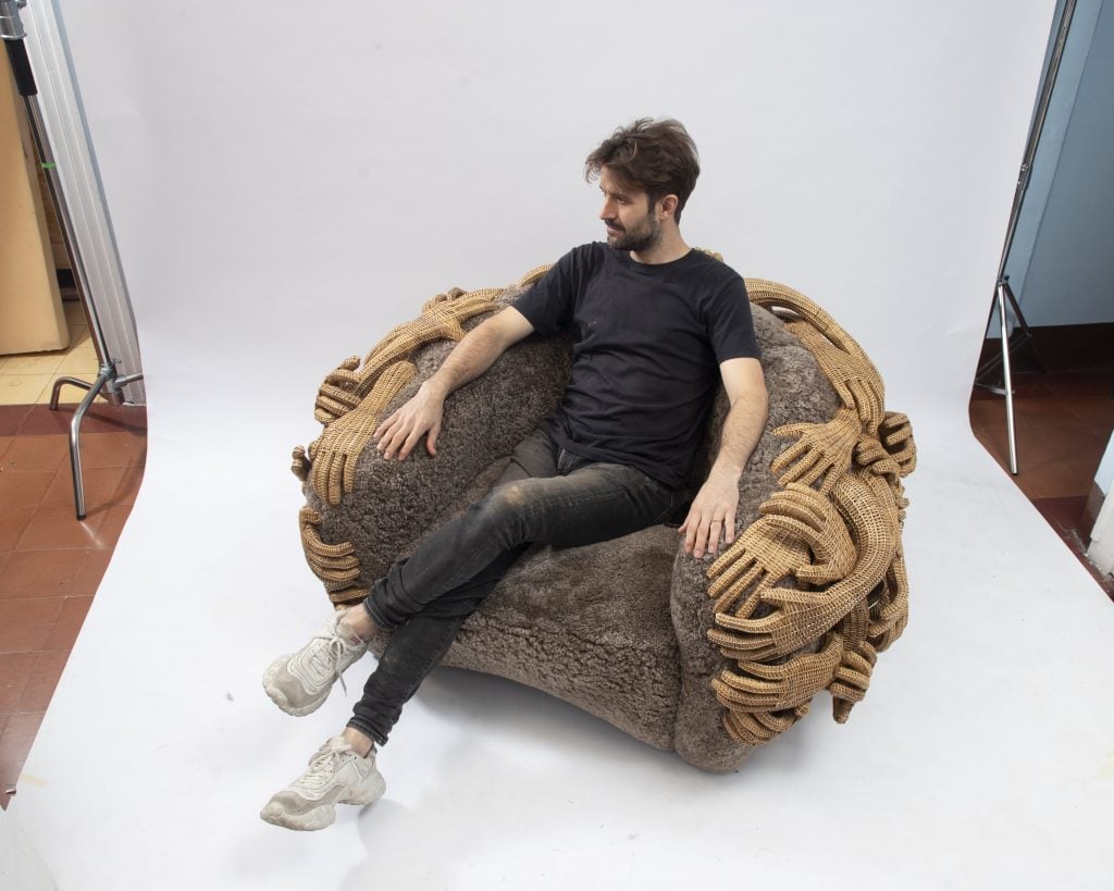 a man sits on a club chair made of a fuzzy seat and wicker arms 