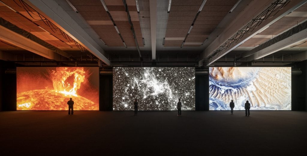 a photo of people standing in a large dark room where 3 large screens show bright images of stars 