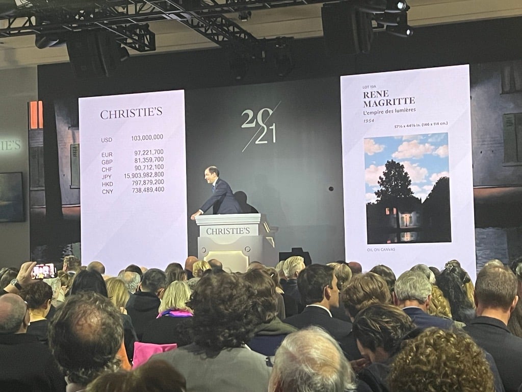 an auction room with a screen showing Rene Magritte's Empire of Light painting