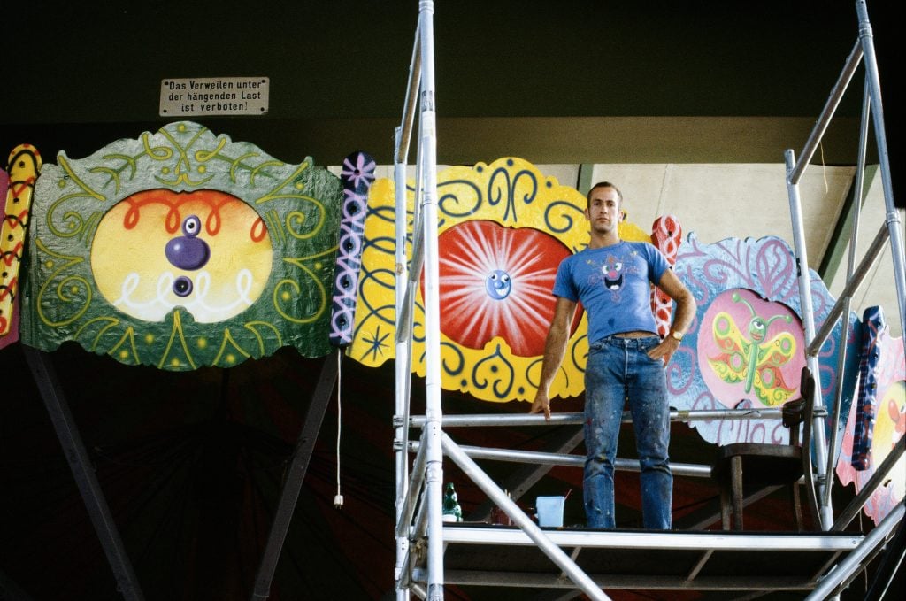 a man stands in jeans and a tight t-shirt on top of scaffolding in front of a painting in progress 