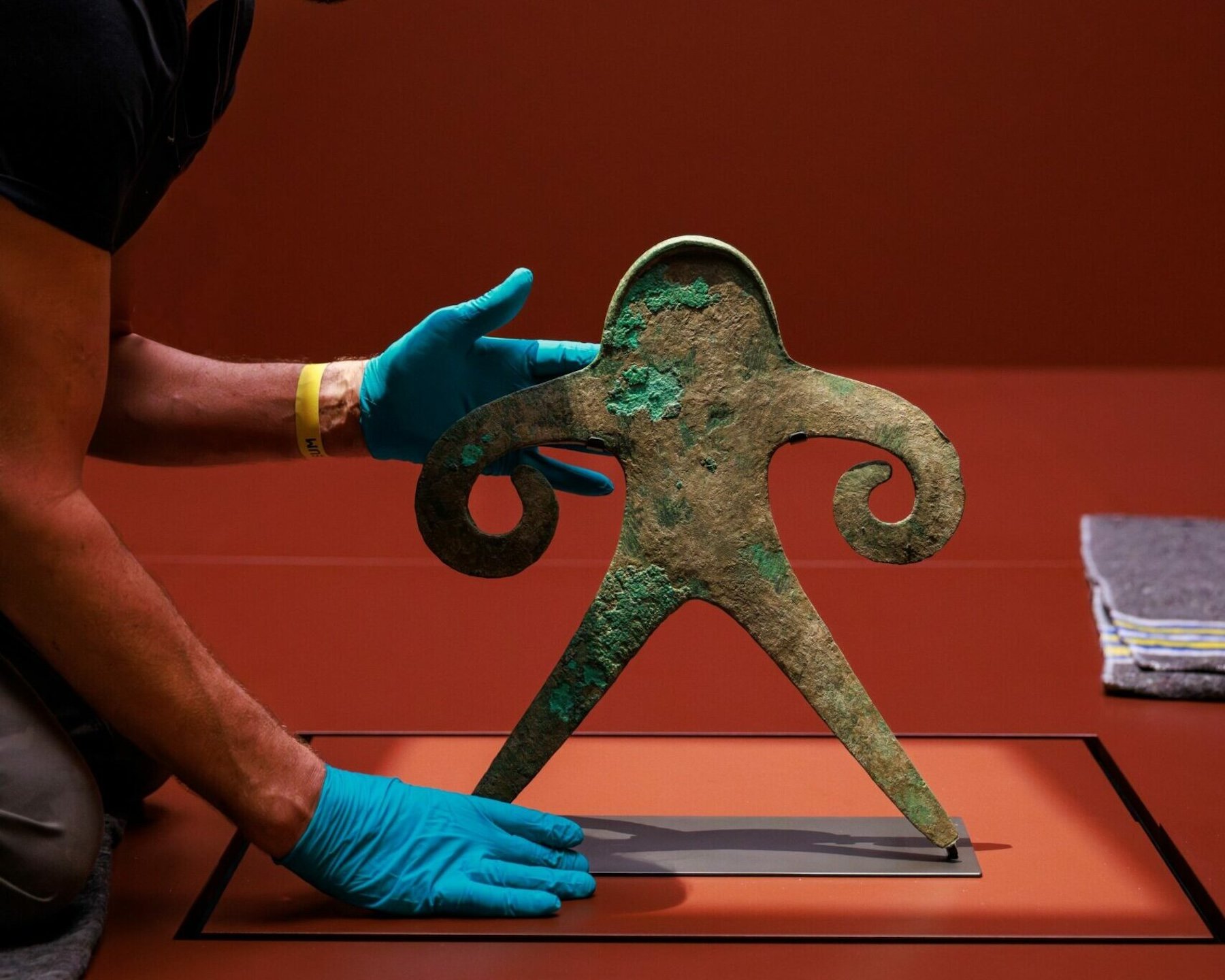 A museum staff member wearing blue gloves carefully places an ancient bronze anthropomorphic figure on display against a red backdrop.