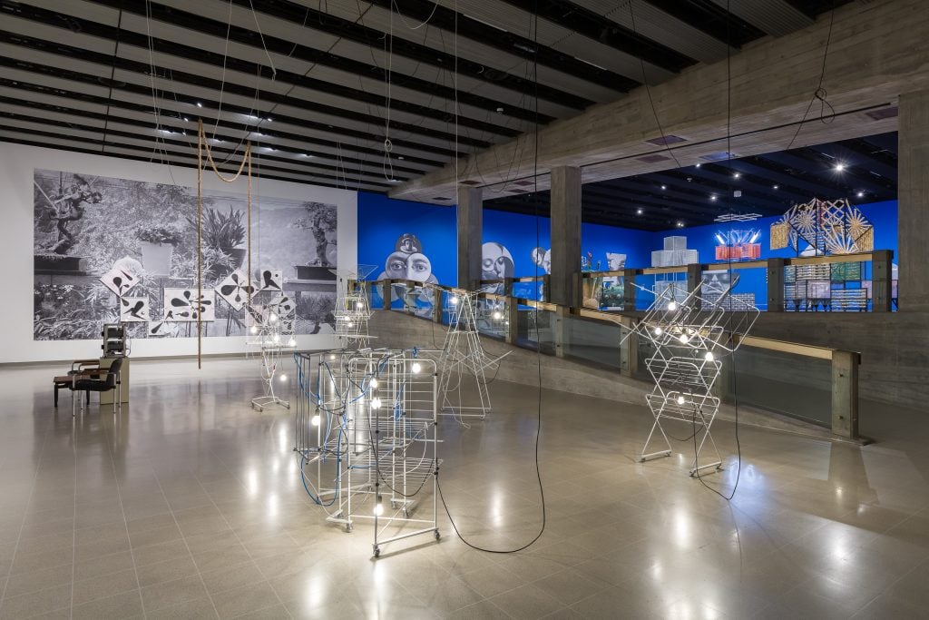  Exhibition space with wire sculptures illuminated by hanging light bulbs, large black-and-white photo mural, blue-painted walls, and various abstract art installations.