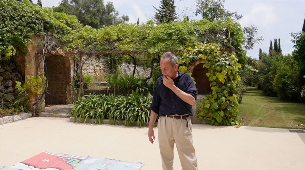 Jean Charles Blais in a blue shirt and beige trousers stood outside of his studio with folliage.