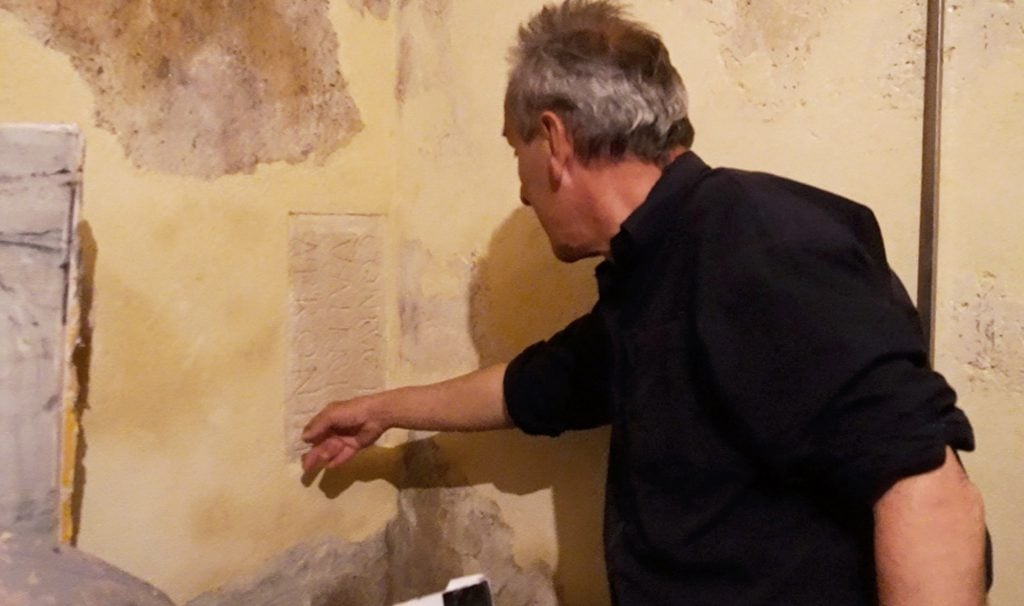 Jean Charles Blais pointing to a Roman inscribed stone which is revealed beneath his studio wall.